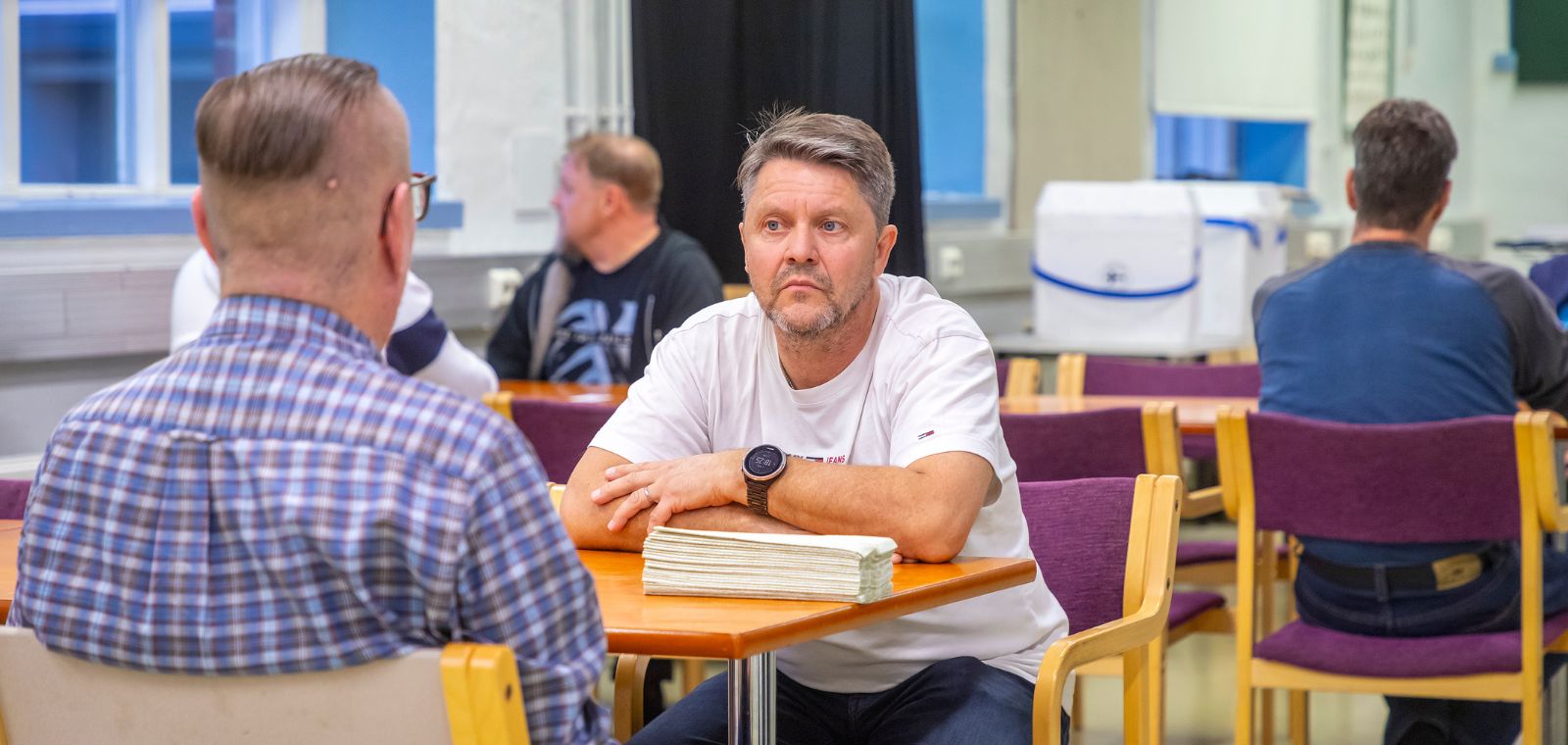 Kaksi miestä istuu toisiaan vastapäätä ja keskustelee, pöydällä iso pino paperisia käsipyyhkeitä.