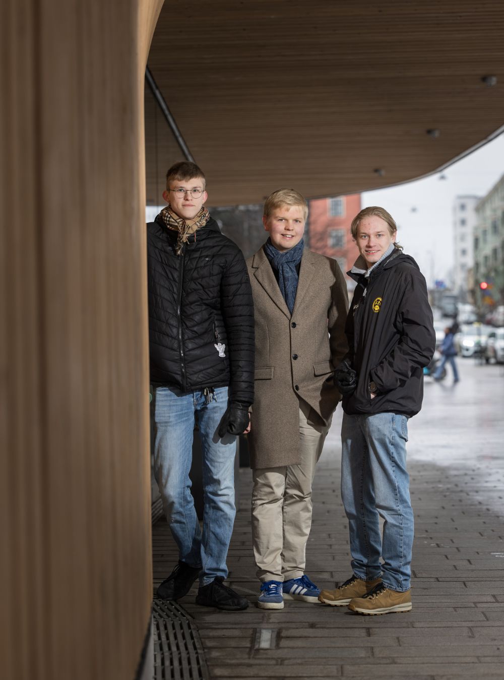 Kolme nuorta miestä seisoo sateelta suojassa Kauppatorin paviljongin katoksen alla.