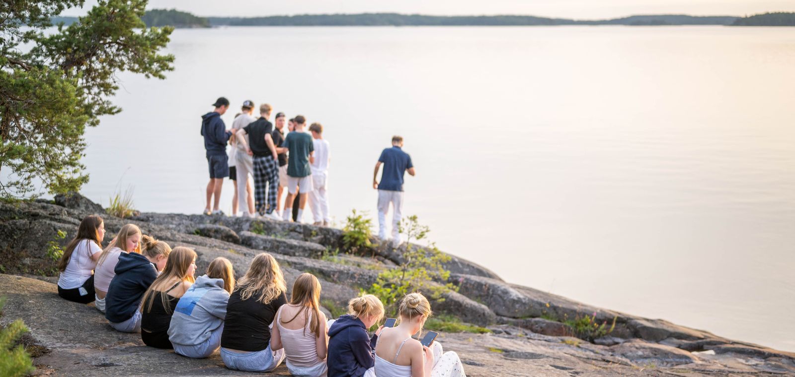 Joukko rippikoululaisia istuu iltaa rantakalliolla.